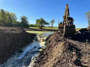 Excavation process for pond renovation in Grain Valley, MO, with a heavy-duty excavator reshaping the landscape and improving water flow for optimal pond management by Bulletproof Pond and Lake. - pond drainage repair